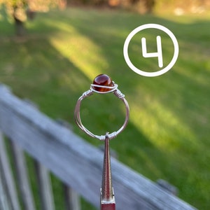 K&ouml;nnte beinhalten: Ein Ring aus silbernem Draht mit einem braunen Achat-Stein. Der Ring wird von einer Zange gehalten.