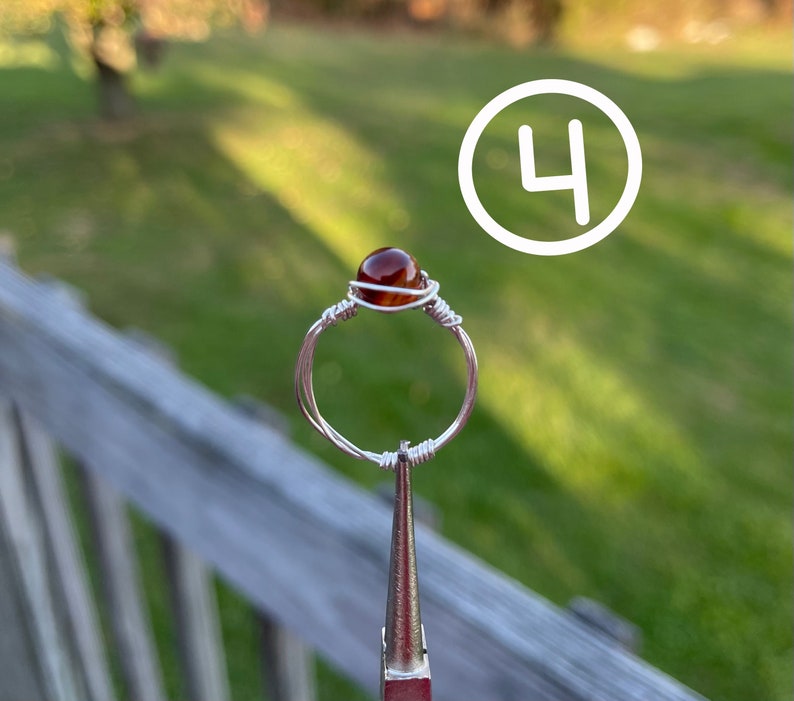 K&ouml;nnte beinhalten: Ein Ring aus silbernem Draht mit einem braunen Achat-Stein. Der Ring wird von einer Zange gehalten.