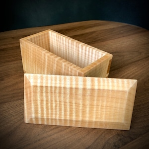 May include: A wooden box with a lid, made of light wood with a distinctive grain pattern. The box is open, revealing the interior.
