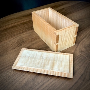 May include: A wooden box with a lid, made of light wood with a natural grain pattern. The box is open, revealing the empty interior. The lid is separate and lying next to the box.