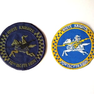 May include: Two embroidered patches with a white knight riding a horse. The patches are circular and have a yellow border with a checkered pattern. The text "White Knights 70th Tac Ftr Sdn" is embroidered on both patches. One patch has a blue background and the other has a dark blue background.
