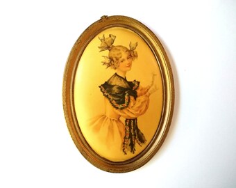 May include: An oval framed portrait of a woman in a yellow dress with a green and black lace collar. She is wearing a white headband with two bows and is holding a white object in her right hand.