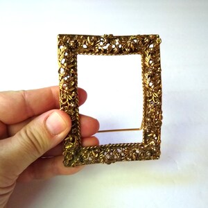 May include: A gold-toned metal picture frame with an ornate design. The frame has a stand on the back and is perfect for displaying a small photo or artwork.