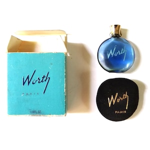 May include: A small blue glass bottle of Worth perfume with a gold cap. The bottle is sitting on a black velvet cushion with the words "Worth Paris" printed in gold. The bottle is also in a light blue and white box with the words "Worth Paris" printed in black.