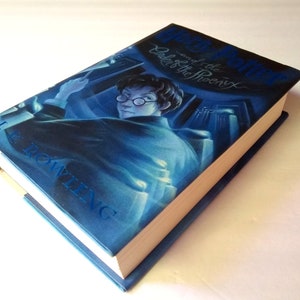 May include: A hardcover book titled "Harry Potter and the Order of the Phoenix" by J.K. Rowling. The cover features a blue-toned illustration of a young man with glasses. The book's spine and edges are blue and the pages are cream-colored.