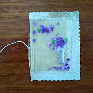 May include: Vintage greeting card with a floral design. The card features a vase of purple flowers and the words "All Loving Greetings". The card has a decorative, scalloped edge and a white string.