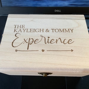 May include: A wooden box with a hinged lid and a metal clasp. The box is engraved with the text "The Kayleigh & Tommy Experience" and an arrow with a heart.