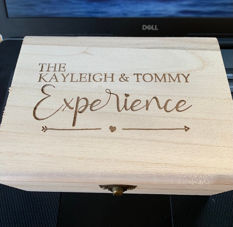 May include: A wooden box with a hinged lid and a metal clasp. The box is engraved with the text "The Kayleigh & Tommy Experience" and an arrow with a heart.