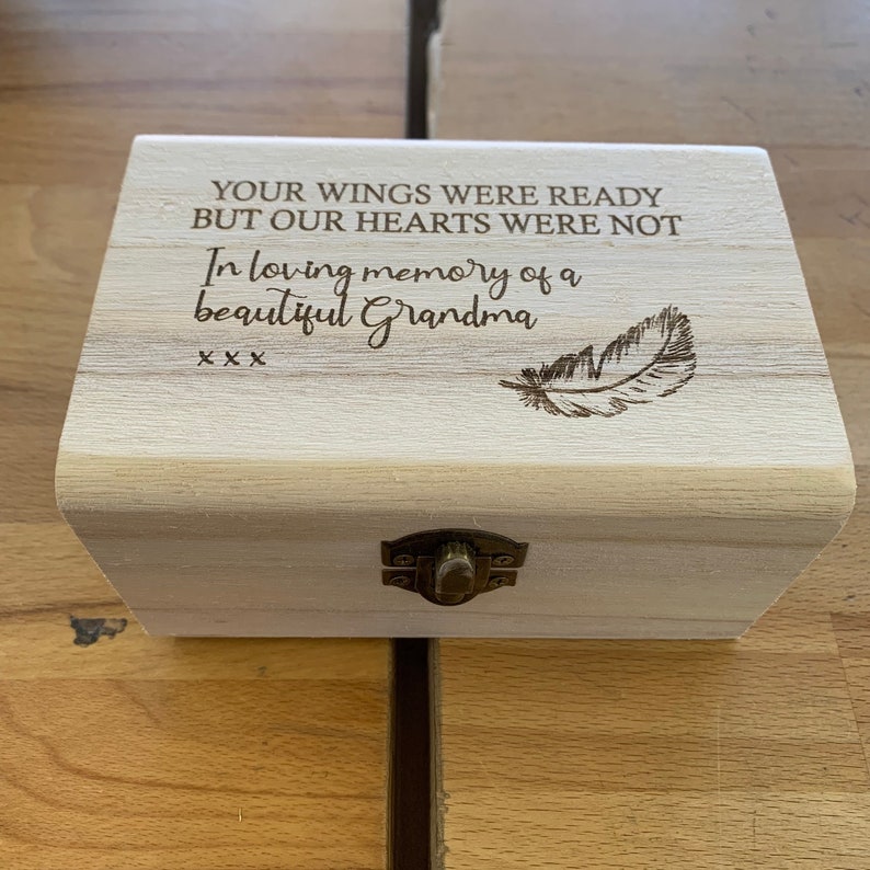 May include: A wooden box with a hinged lid and a brass latch. The box is engraved with the words "Your wings were ready but our hearts were not. In loving memory of a beautiful Grandma. XXX" and a feather design.