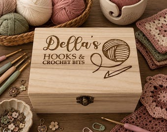 Puede incluir: Una caja de almacenamiento rectangular de madera clara con un cierre de metal, que presenta el texto grabado con láser "Della's HOOKS & CROCHET BITS" y un gráfico de un ovillo de lana y un ganchillo en la tapa, rodeada de varios suministros de ganchillo.