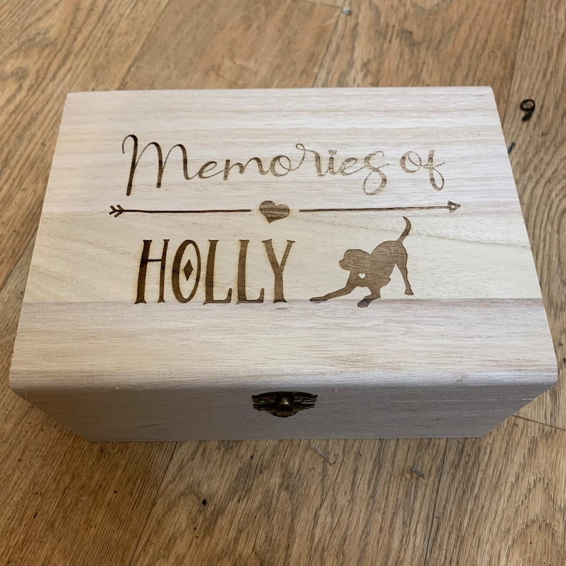 May include: A light wood box with a hinged lid and a brass latch. The box is engraved with the words "Memories of HOLLY" and a silhouette of a dog.