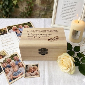May include: A light-coloured wooden keepsake box with a metal latch, engraved with the text "YOUR WINGS WERE READY BUT OUR HEARTS WERE NOT" and "In loving memory of a beautiful Grandma" alongside a feather illustration, surrounded by photos and a candle.