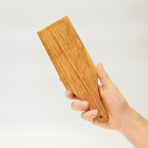 Puede incluir: Una espátula de madera con un acabado marrón claro y un pequeño agujero en el mango.