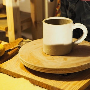 Peut inclure: Une tasse en c&eacute;ramique blanche avec un gla&ccedil;age brun sur le fond est pos&eacute;e sur un tour de potier en bois.