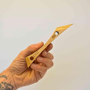 Può includere: Uno strumento in legno con una punta appuntita e un manico. Il manico ha due fori con il testo "CO2" e "A&B" inciso su di esso.