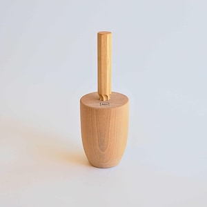 Puede incluir: Un mortero de madera clara con una base cilíndrica y un mango vertical. La base es redondeada y el mango tiene forma rectangular recta. La veta de la madera es visible. Un pequeño logotipo cuadrado está en la base.