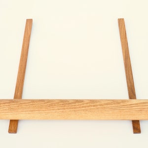 May include: A wooden rolling pin resting on two wooden supports. The rolling pin is light brown and has a natural wood grain. The supports are dark brown and are positioned parallel to each other.