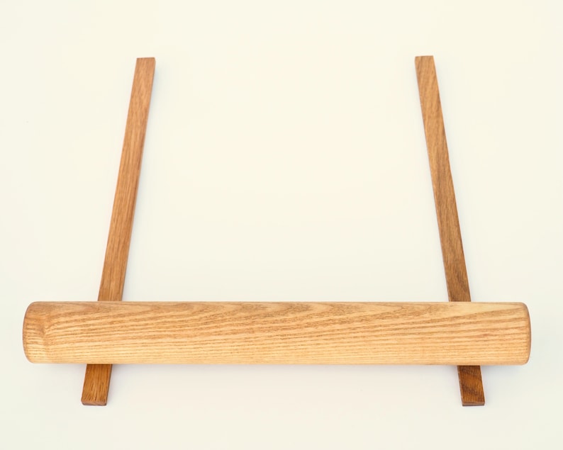 May include: A wooden rolling pin resting on two wooden supports. The rolling pin is light brown and has a natural wood grain. The supports are dark brown and are positioned parallel to each other.