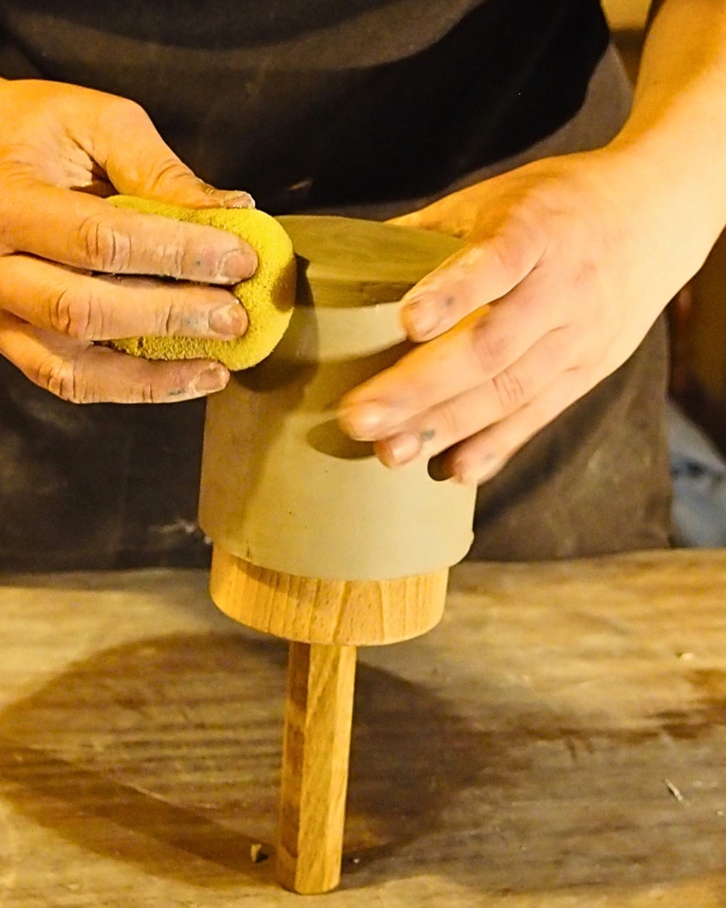 Peut inclure: Une personne lisse le dessus d'un pot en argile avec une &eacute;ponge jaune. Le pot est pos&eacute; sur une base en bois.