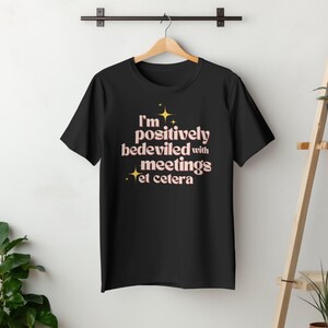 May include: Black t-shirt with the text "I'm positively bedeviled with meetings et cetera" in a light pink font with gold star accents. The shirt is hanging on a wooden hanger against a white wall.