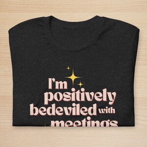 May include: Dark gray t-shirt with the text "I'm positively bedeviled with meetings" in a retro font. The text is in a light pink color with a yellow starburst graphic above the text. The shirt is folded on a light wood surface.