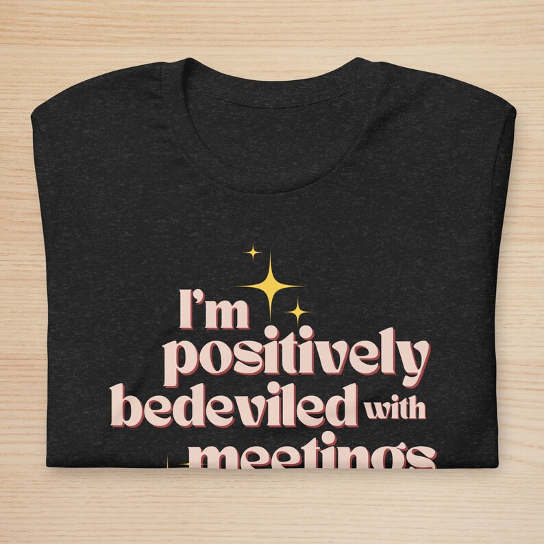 May include: Dark gray t-shirt with the text "I'm positively bedeviled with meetings" in a retro font. The text is in a light pink color with a yellow starburst graphic above the text. The shirt is folded on a light wood surface.