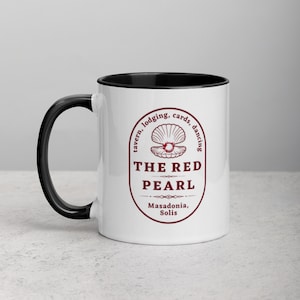 Close-up studio shot of a white ceramic mug with a black handle and interior. The mug is oriented with the handle facing towards the left. Centered on the mug is a maroon-colored oval logo containing "The Red Pearl" in bold sans-serif letters. Above this title is shell graphic, around logo text include tavern, lodging, cards, dancing, and location "Masadonia, Solis". The image has balanced lighting and is placed against a gradient soft gray background.