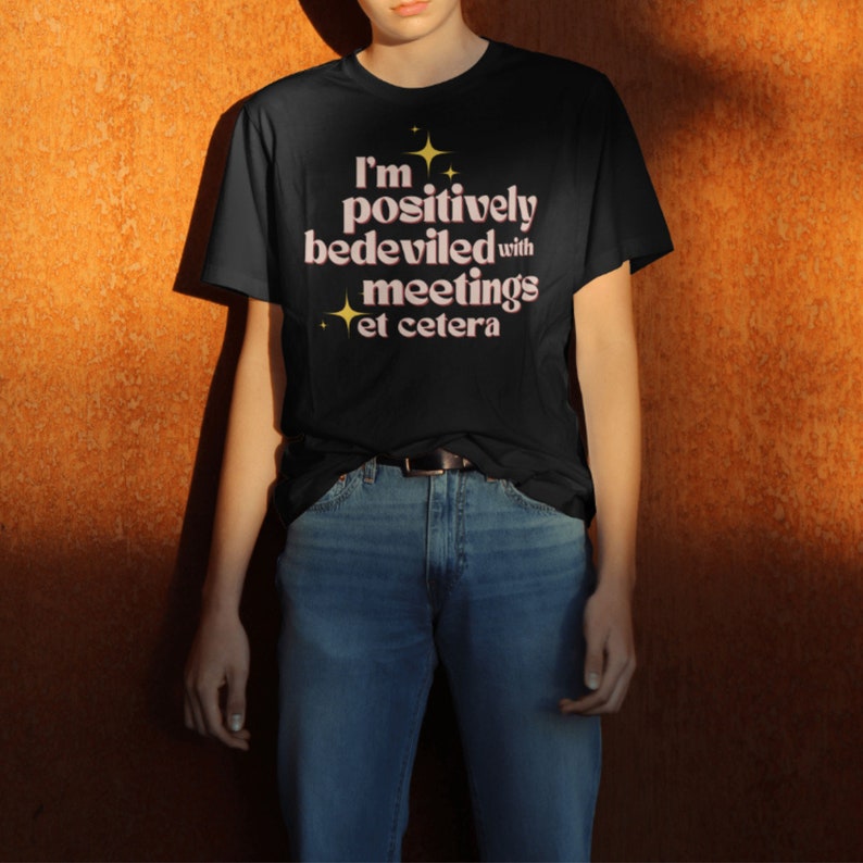 May include: Black t-shirt with the text "I'm positively bedeviled with meetings et cetera" in a retro font. The text is in pink with gold star accents. The person is wearing blue jeans and a black belt.