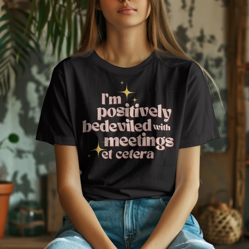 A young woman wears a black t-shirt featuring Moira Rose from Schitt's Creek iconic quote "I am positively bedeviled with meetings, et cetera" in a bold font in light pink. There are yellow stars at the top and bottom left of the quote giving it a bit of a sparkle. The design is simple, capturing the essence of the TV series in a refined and artistic style. The woman sits in a cozy indoor space with plants and rustic decor, dressed casually in blue jeans, creating a warm, artistic atmosphere.