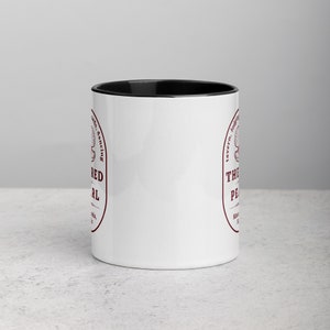 Close-up studio shot of a white ceramic mug with a black handle and interior. The mug is oriented with the handle facing towards the left. Centered on the mug is a maroon-colored oval logo containing "The Red Pearl" in bold sans-serif letters. Above this title is shell graphic, around logo text include tavern, lodging, cards, dancing, and location "Masadonia, Solis". The image has balanced lighting and is placed against a gradient soft gray background.