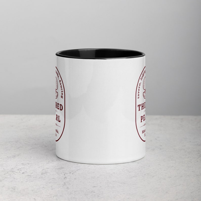 Close-up studio shot of a white ceramic mug with a black handle and interior. The mug is oriented with the handle facing towards the left. Centered on the mug is a maroon-colored oval logo containing "The Red Pearl" in bold sans-serif letters. Above this title is shell graphic, around logo text include tavern, lodging, cards, dancing, and location "Masadonia, Solis". The image has balanced lighting and is placed against a gradient soft gray background.