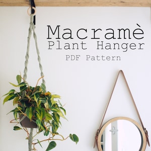 Può includere: Un portavasi in macramè con un cordoncino verde e un anello di legno in cima. Il portavasi tiene una pianta in vaso con foglie verdi. Il testo "Macrame Plant Hanger PDF Pattern Digital download" è scritto sotto il portavasi.