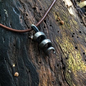 Puede incluir: Un colgante de metal gris oscuro con forma de espiral o bobina apretada, suspendido de un cordón de cuero marrón liso. El colgante tiene una apariencia texturizada forjada a mano con un acabado mate, mostrando un diseño simple y curvado.
