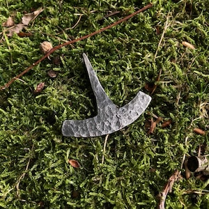 Könnte beinhalten: Ein kleiner, handgeschmiedeter Metall-Axtkopf mit strukturierter Oberfläche. Der Axtkopf ist grau und liegt auf einem Bett aus leuchtend grüner Moos. Der Axtkopf hat eine gebogene Klinge und eine spitze Spitze.