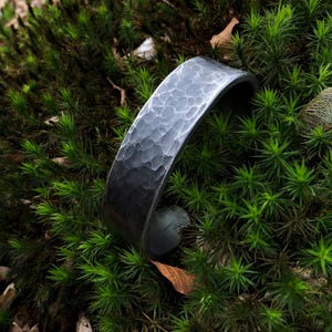 Könnte beinhalten: Ein dunkelgraues, gehämmertes Metallarmband mit strukturierter Oberfläche, das auf grünem Moos liegt. Das Armband ist ein schlichtes, aber elegantes Schmuckstück.