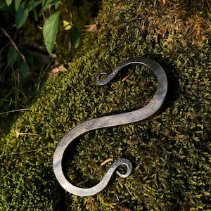 May include: A hand-forged S-shaped metal hook with a dark, textured finish. The hook features curved, scrolled ends that taper to points, creating a continuous, flowing form. The metal surface displays a hammered, matte grey appearance.