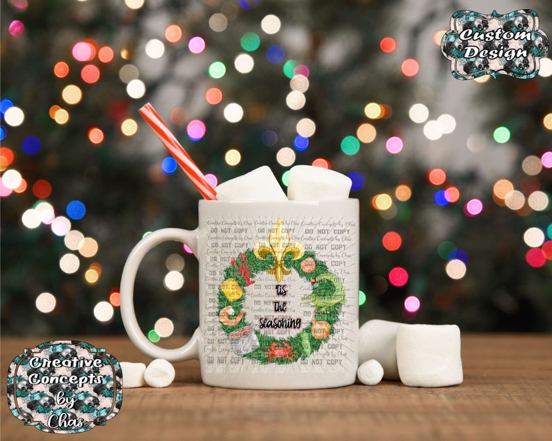 May include: A white ceramic mug filled with marshmallows and a red and white striped straw. The mug features a green wreath graphic with a gold fleur-de-lis, seafood illustrations, and the black text 'tis the seasoning' in the center.