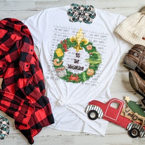 May include: A white t-shirt featuring a graphic of a green wreath decorated with seafood and a gold fleur-de-lis, with the text 'tis the seasoning' in the center. The shirt is displayed with a red and black plaid flannel shirt, a cream beanie, and brown boots.