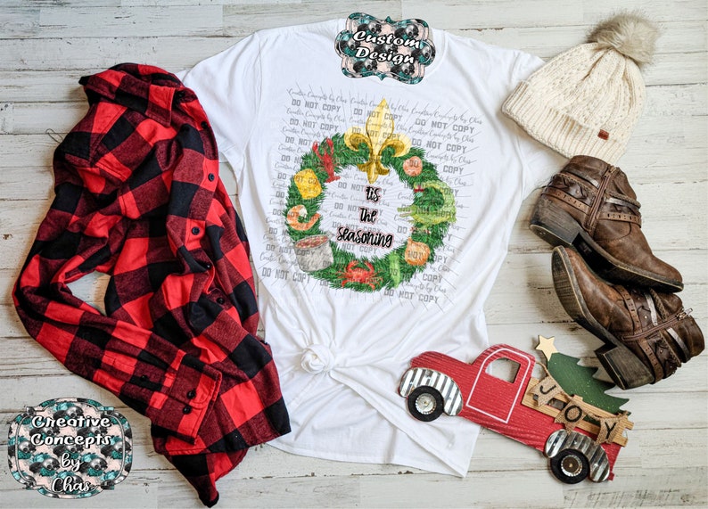May include: A white t-shirt featuring a graphic of a green wreath decorated with seafood and a gold fleur-de-lis, with the text 'tis the seasoning' in the center. The shirt is displayed with a red and black plaid flannel shirt, a cream beanie, and brown boots.