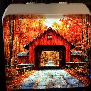 May include: A night light featuring a vibrant autumn scene. A red covered bridge is the focal point, surrounded by trees with orange and yellow foliage. The bridge spans a road, creating a picturesque landscape.