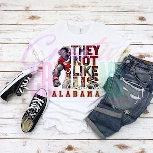 May include: White t-shirt with a red, white, and black graphic of an elephant wearing a football uniform. The text "They Not Like Us Crimson Tide Alabama" is printed on the shirt.