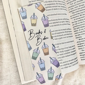 May include: A bookmark with illustrations of bubble tea in pastel colours: blue, green, purple, and beige. The text "Books & Boba" is written in a stylish font. The bookmark is inside an open book.