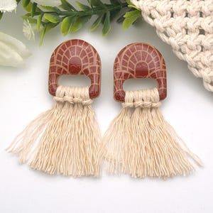 May include: A pair of earrings featuring a semi-circular terracotta-coloured component with a white grid pattern. Each piece is attached to a long, cream-coloured cotton cord fringe that hangs downward in a series of vertical, thin strands.