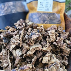 May include: A pile of dried, brown, irregular-shaped beef lung dog treats in the foreground. In the background, two stand-up pouches are visible, one with a tan label reading Handcrafted Dog Treats and Beef Lung, and one black pouch.