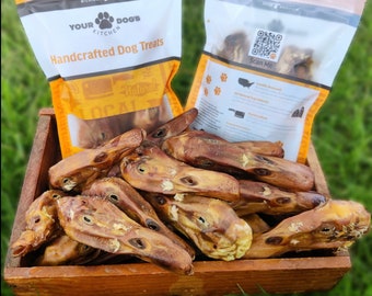 Puede incluir: Una caja de madera llena de cabezas de pato secas para mascotas, colocada frente a dos bolsas de plástico blancas y naranjas etiquetadas Your Dog's Kitchen Handcrafted Dog Treats con un código QR y texto sobre ingredientes locales.