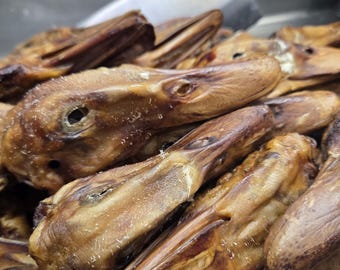 Puede incluir: Un montón de varias cabezas de pato ahumadas con una textura de piel marrón y seca y picos visibles. Un cuchillo de metal con mango de madera oscura se encuentra en el fondo, parcialmente visible detrás de la pila de cabezas de pato.