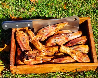 Puede incluir: Una caja de madera rectangular llena de varias cabezas de pato secas de color marrón anaranjado. Un cuchillo de carnicero grande de acero inoxidable con mango negro y tres remaches plateados descansa horizontalmente sobre la caja.