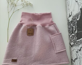 May include: A light pink skirt with a high waistband and a small pocket. The skirt has a tag attached with the brand name. The skirt is made of a soft, textured material. A sprig of white flowers is in the background.