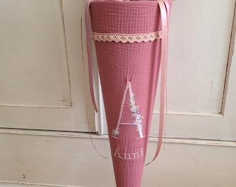 May include: A dusty rose-colored cone-shaped school bag with a gathered top and ribbon ties. The bag features a white embroidered letter "A" with floral accents and the name "Anni" below. A delicate lace trim adorns the top.