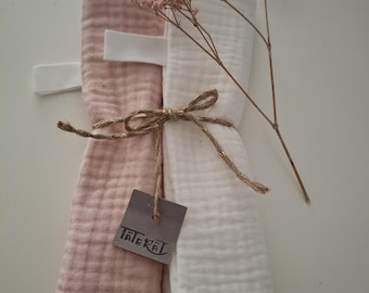 Puede incluir: Un paquete de dos muselinas, una rosa rubor y otra blanca, atadas con hilo. Una pequeña etiqueta con el texto "TALEKAL" cuelga del hilo. Flores secas se colocan encima del paquete.